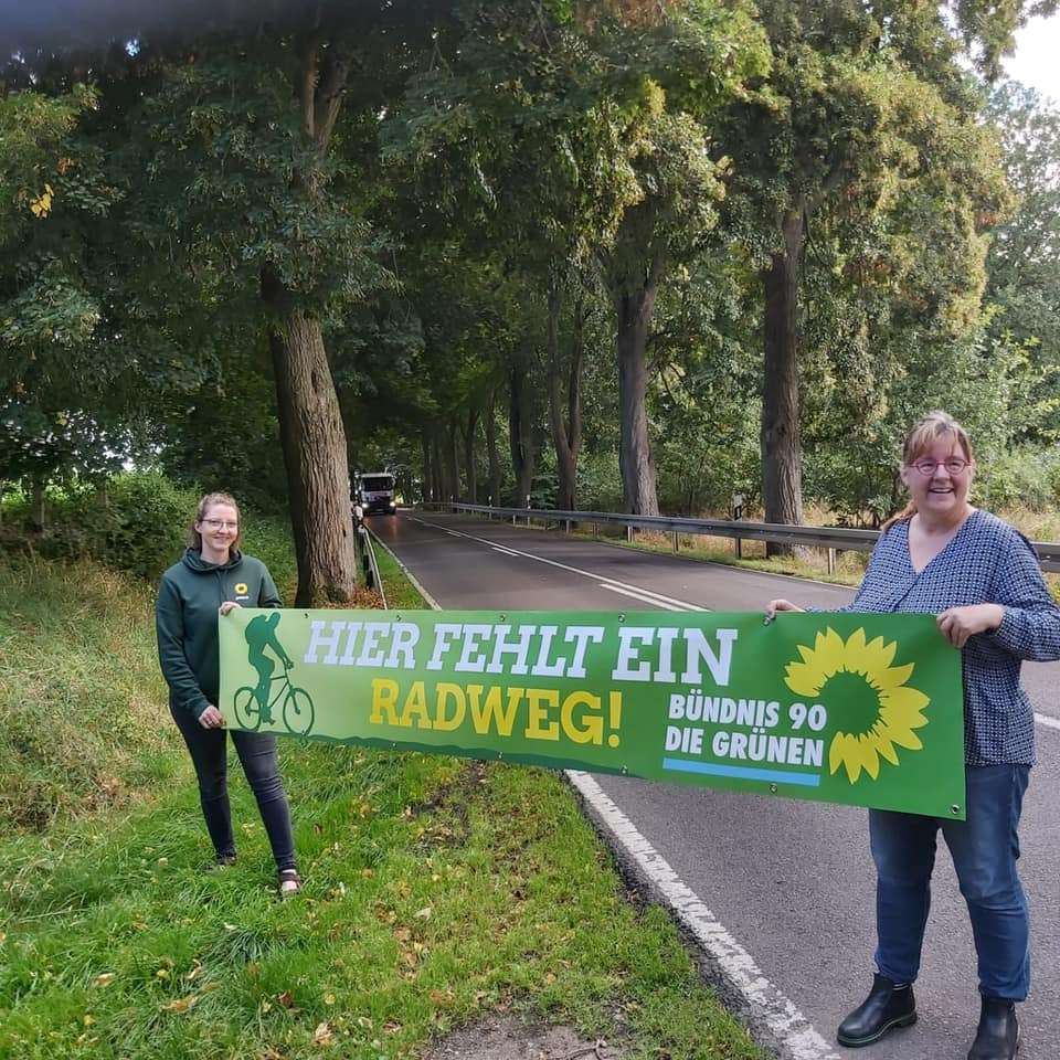 Friederike Fiß und Jutta Wegner stehen am Straßenrand und halten ein Banner mit der Aufschrift "Hier fehlt ein Radweg".